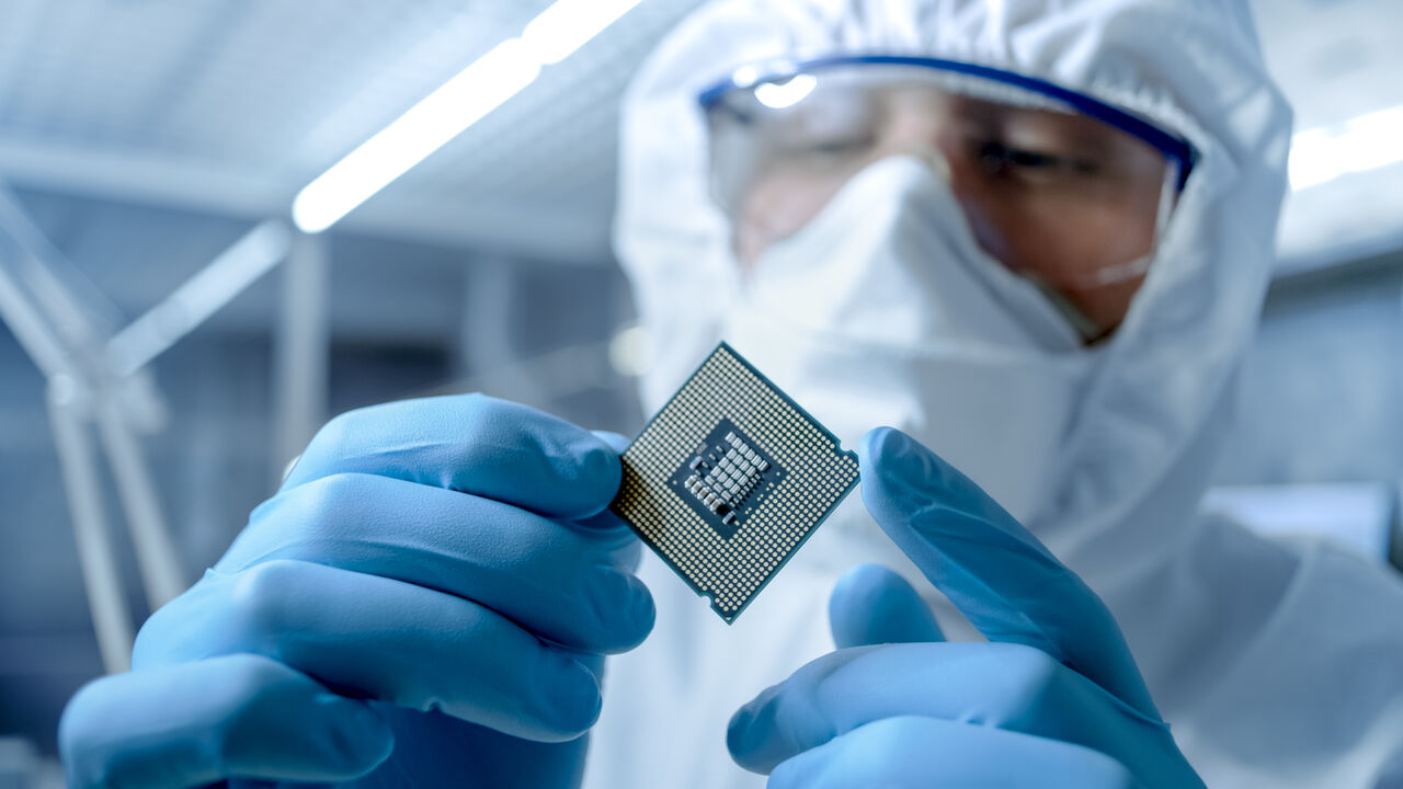 In Ultra Modern Electronic Manufacturing Factory Design Engineer in Sterile Coverall Holds Microchip with Gloves and Examines it.